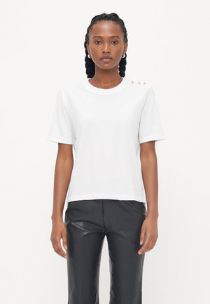 T-shirt en coton blanc à manches courtes, col rond, et trois boutons décoratifs sur l'épaule gauche. Porté avec un pantalon en cuir noir.