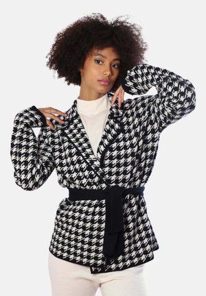 Donna con capelli ricci che indossa un cardigan con cintura e motivo pied de poule nero e bianco sopra un dolcevita bianco e pantaloni, posando con le mani vicino al viso.