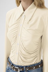 Kremfarget bluse med lange ermer og knapper foran, med detaljer av rynking på sidene og en spiss krage, kombinert med blå jeans og et brunt belte.