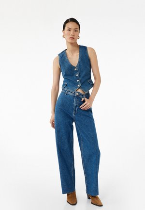 Kamizelka dżinsowa z dekoltem w kształcie litery V i guzikami, zestawiona z szerokimi dżinsami. Obie części są w jednolitym ciemnoniebieskim odcieniu, z minimalnymi przeszyciami.