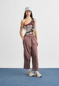 Débardeur asymétrique avec un motif graphique en noir, jaune et rouge ; pantalon ample marron ; casquette gris clair ; baskets grises et blanches.