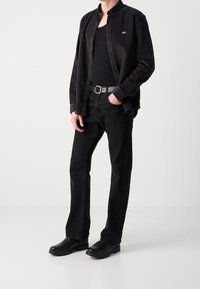 Camisa de denim negra sobre una camiseta negra de tirantes, combinada con jeans negros y zapatillas negras sin cordones. Presenta un pequeño logo en la camisa.