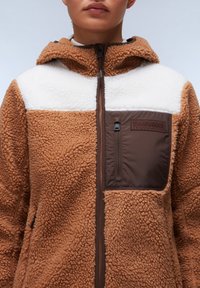 Bruine fleece jack met een witte bovenpaneel. Heeft een ritszak op de linker borst, textuurstof en een capuchon.