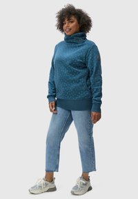 Felpa blu con motivo e colletto a imbuto, arricchita da dettagli con bottoni, abbinata a jeans azzurri tagliati e sneaker grigie.