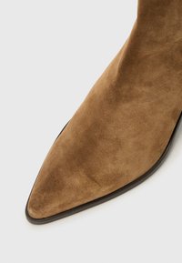 Gros plan sur une botte en daim marron à bout pointu, montrant sa texture souple et son design épuré sur un fond blanc.