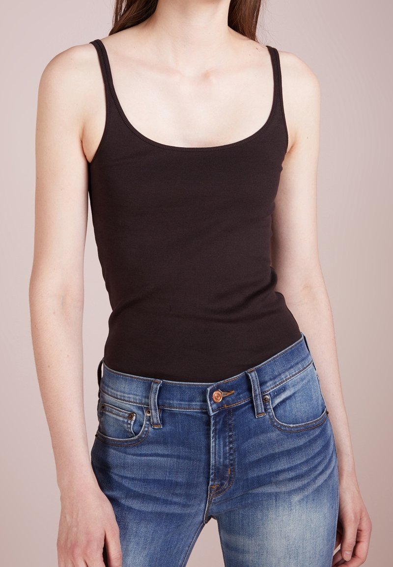 Femme portant un débardeur noir près du corps et un jean bleu, debout devant un fond neutre uni.