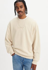 Beige sweatshirt laget av mykt stoff, med et teksturert "Levi's"-logo på brystet, avslappet passform, senket skuldre og mansjetter.