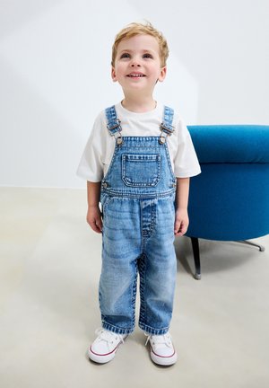 Blue denim overalls with adjustable straps, front pocket, loose-fitting legs, paired with a white t-shirt and white sneakers with red accents.