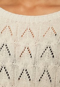 Ljust beige stickad tröja med ett geometriskt mönster med öppna stygn, vertikala och diagonala linjer samt subtila svarta accenter.