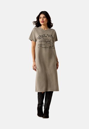 Frau trägt ein beiges Midikleid im T-Shirt-Stil mit dem Text "Wild West" und einer Stier-Grafik, kombiniert mit schwarzen, bestickten kniehohen Stiefeln.