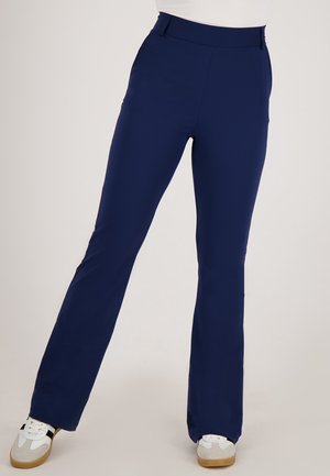 Persoon in marineblauwe rechte broek en witte sneakers, staande met de benen iets gekruist tegen een effen achtergrond.