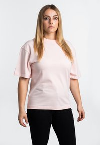 Lys rosa kortermet t-skjorte laget av bomull, med rund hals og en avslappet passform. Modellen står med svarte bukser foran en nøytral bakgrunn.