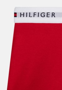 Piros pamut szövet, fehér rugalmas derékkal, amelyen fekete betűkkel a "HILFIGER" felirat látható, kísérve egy kis, sokszínű logóval.