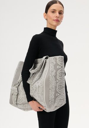 Kobieta w czarnym golfie trzyma dużą szarą torbę na ramię z białym wzorem paisley na jednolitym białym tle.