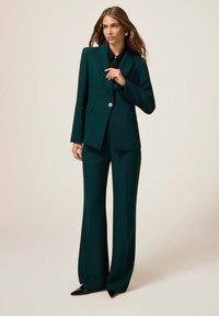 Traje ajustado en color verde azulado con pantalones de pierna ancha y una chaqueta estructurada, que cuenta con un botón único y bolsillos de solapa, combinado con una camisa negra.