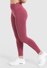 Hoge taille roze leggings van rekbaar materiaal, met een gladde textuur en een enkellange snit, gedragen met witte sportieve schoenen.