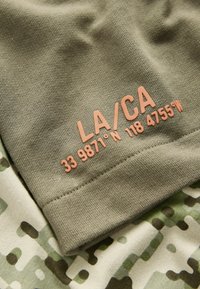 Olivgrüner Stoff mit pinkem Text, der "LA/CA 33 9871°N 118 4755°W" liest. Der Hintergrund zeigt einen beigen Stoff mit einem Tarnmuster.