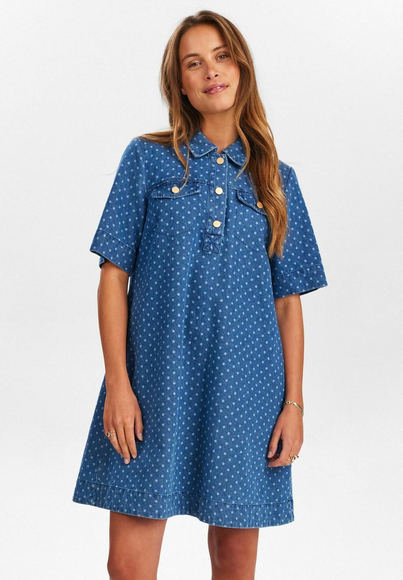 Robe en denim bleu foncé avec des pois blancs, manches courtes, col boutonné et deux poches poitrine. Coupe évasée avec un ourlet au-dessus du genou.