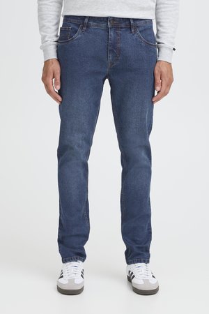 Mann trägt blaue Jeans, weiße Turnschuhe mit schwarzen Streifen und ein hellgraues Sweatshirt, steht vor einem schlichten weißen Hintergrund.