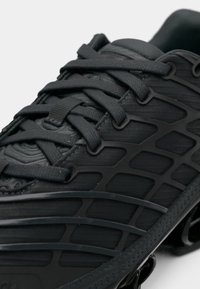 Chaussure de sport noire dotée d'un matériau en mesh, d'un design texturé, de lacets épais et d'accents moulés sur le côté. Semelle extérieure amortissante visible.