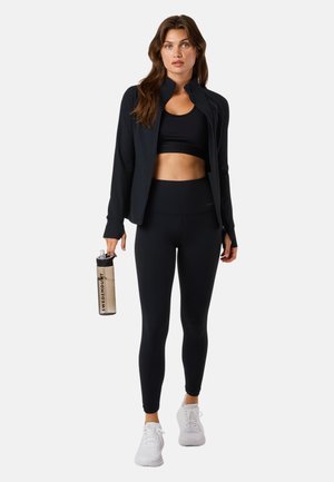 Vrouw die zwarte sportleggings, een crop top en een jas draagt, met een beige waterfles met de tekst "Swedemount" in haar hand, staand tegen een witte achtergrond.