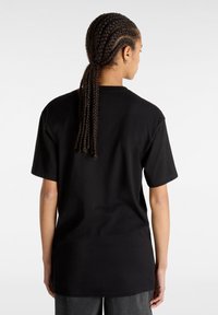 Personne aux cheveux tressés attachés, portant un T-shirt noir uni à manches courtes et un pantalon foncé, vue de dos sur un fond blanc.