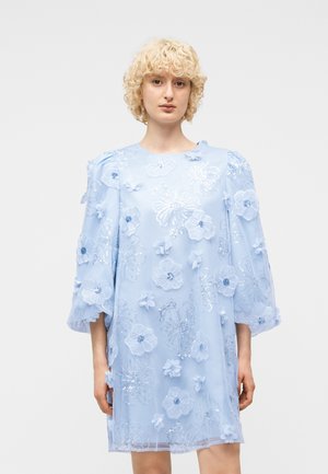 Nainen pukeutuneena vaaleansiniseen mekkoon, jossa on kolmiulotteisia kukka-aplikeerauksia ja paljetteja, väljät kolme neljäsosan hihat ja pyöreä pääntie.