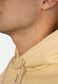 Hoodie beige chiaro con tessuto testurizzato, un cordoncino con accessori dorati e branding ricamato nella zona del colletto.