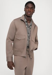 Man wearing beige zip-up jacket with large pockets, beige pants, and a snakeskin patterned shirt underneath, standing against plain background.