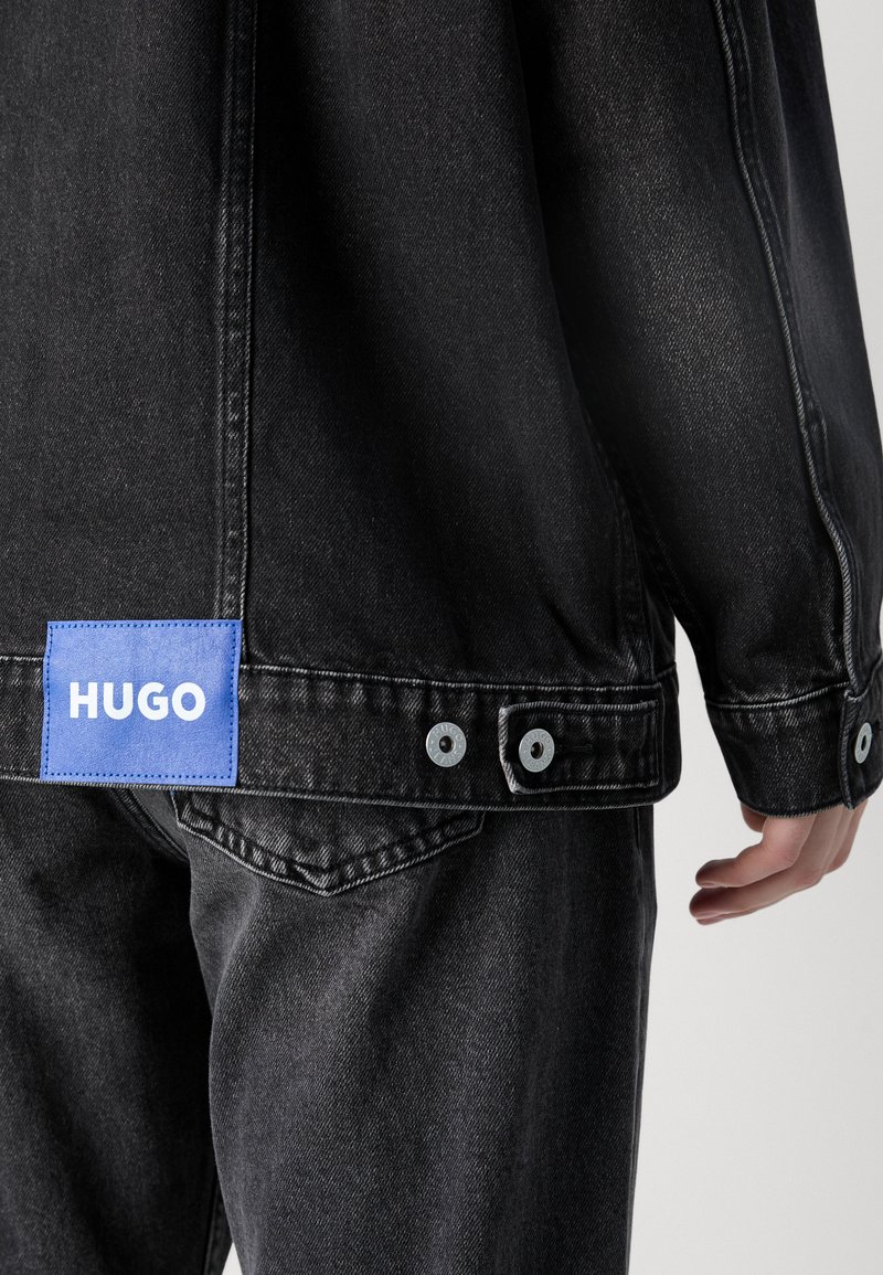 Czarna kurtka dżinsowa z niebieską naszywką marki, srebrnymi zatrzaskami, widocznym przeszyciem, luźnym krojem i teksturowaną powierzchnią.