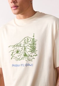 Camiseta de algodón color beige claro con una ilustración de un campamento verde que incluye una tienda de campaña, árboles y montañas, además de texto azul que dice "PHOENIX 112.0740."