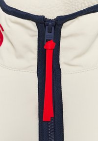 Giacca beige con zip, dotata di un colletto e una zip blu navy, e un tirazip rosso brillante, con un interno in pile per un ulteriore calore.