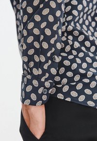 Granatowa koszula w kwiatowy wzór w odcieniach kremu i jasnego brązu, z zapięciem na guzik przy mankietach i gładką teksturą materiału.