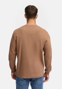 Camisa marrón de manga larga con una tela texturada, cuello redondo y puños acanalados, combinada con vaqueros de un azul claro.