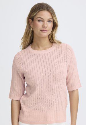 Noor naine õlgadeni blondide juustega, kes kannab rõngaskõrvarõngaid ja heleroosat lühikeste varrukatega kudumitoppi, seisab näoga ettepoole neutraalse ilmega.