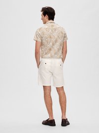 Chemise à manches courtes beige avec un motif de feuilles de palmier, associée à un short blanc. Des chaussures brunes complètent l'ensemble. Design simple et décontracté.