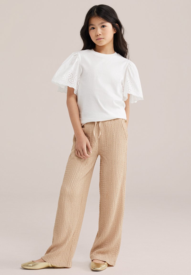 Djevojka stoji noseći bijelu majicu s čipkastim lepršavim rukavima, široke hlače bež teksture i zlatne ravne cipele na neutralnoj pozadini.