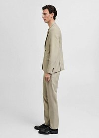 Veste et pantalon de costume beige sur mesure, tissu lisse, fermeture à un bouton, éclat subtil, associés à des chaussures en cuir noires.