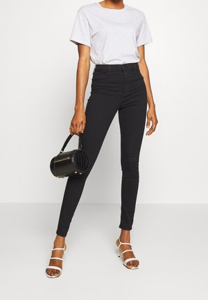 Jeans ajustados de cintura alta en negro, confeccionados en denim, combinados con una camiseta gris clara y tacones blancos de tiras. Sosteniendo un bolso negro redondo.