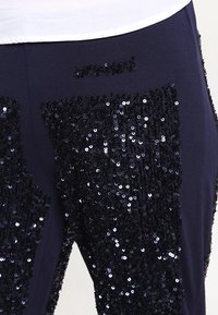 Marinblå byxor med ett paljettmönster som täcker framsidan, med en slät textur och en figurnära design, lämpliga för både avslappnade och mer eleganta tillfällen.