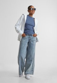 Felpa bianca con cappuccio, canotta azzurra a coste, jeans oversize blu chiaro, sneakers bianche, occhiali da sole neri, accessori in argento, modella sorridente.