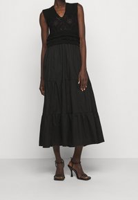 Frau in einem ärmellosen schwarzen Stricktop mit Spitzenmustern, einem gestuften schwarzen Midikleid und schwarzen High-Heels.