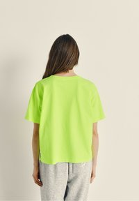 Neon žalia trumpa rankovė T-shirt su laisvu siluetu, apvaliu iškirpimu ir šoninėmis skeltukais. Audinys atrodo lygus ir lengvas.