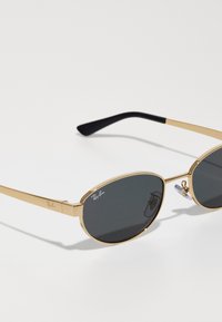 Ochelari de soare ovali cu lentile gri închis, ramă subțire din metal auriu și brațe negre. Prezintă logo-ul Ray-Ban pe ramă.