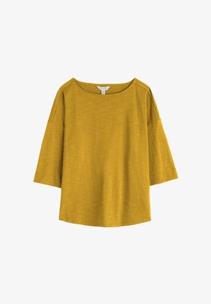 Mosterdgele top met losse pasvorm, brede mouwen tot aan de ellebogen en een ronde halslijn op een witte achtergrond.