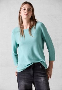 Türkiser strukturierter Pullover mit Dreiviertelärmeln, kombiniert mit dunkelblauen Jeans. Mit Rundhalsausschnitt und lockerer Passform.