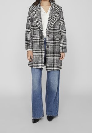 Femme portant un manteau à carreaux noir et blanc, un chemisier blanc, un jean bleu à jambes larges et des chaussures noires à bouts pointus, debout sur fond gris.
