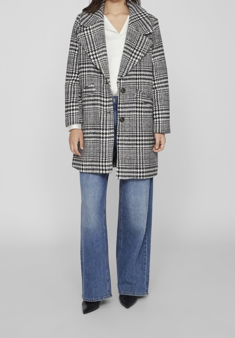 Femme portant un manteau à carreaux noir et blanc, un chemisier blanc, un jean bleu à jambes larges et des chaussures noires à bouts pointus, debout sur fond gris.