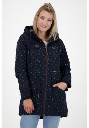Eine lächelnde Frau trägt eine navyblaue gepunktete Kapuzenjacke, die zugezippt ist und ihre Hände in den Taschen hat, kombiniert mit hellblauen Jeans.