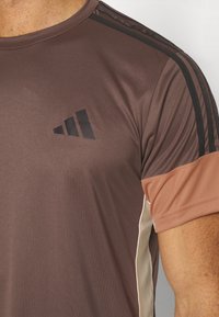 Camiseta atlética marrón hecha de una tela suave, con rayas negras en los hombros y un logo en el pecho. Mangas cortas con acentos beige.